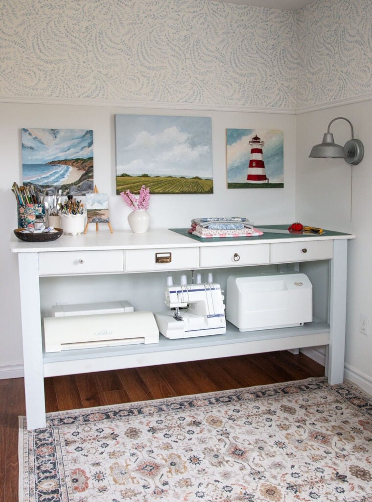 A DIY craft table with storage set against a white wall with blue leafy patterned wallpaper above a chair rail. The table is painted white with a light gray shelf below, holding a cutting machine, a serger sewing machine, and another crafting machine. On top of the table, there are organized supplies including jars of paintbrushes, a small vase of pink flowers, a stack of folded fabrics, and a cutting mat. Three coastal-themed paintings hang above the table, and a gray industrial-style wall sconce is mounted to the right. A vintage-style patterned rug covers the wood floor beneath the table.