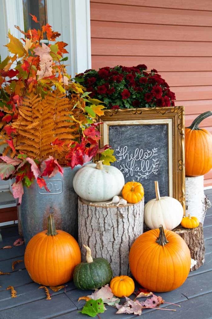 Hello Fall sign on a fall front porch. Fall front porch decorated with vibrant orange mums, small white pumpkins, and colorful autumn leaves in rustic baskets