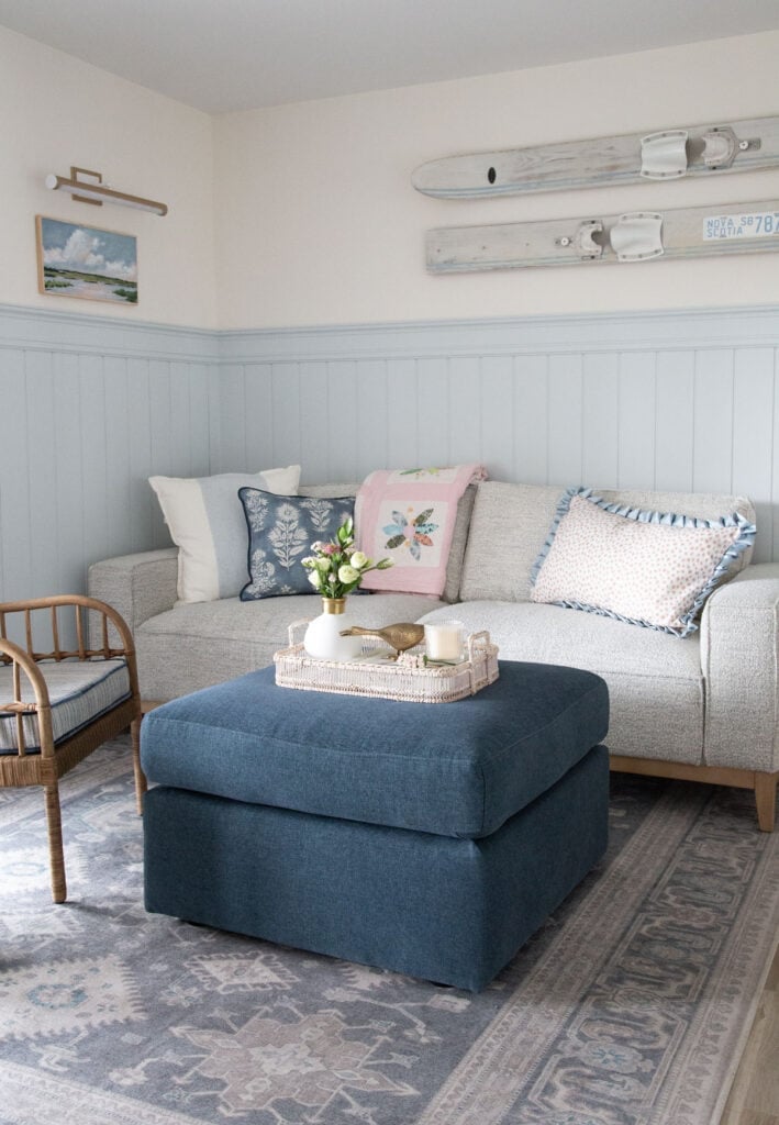 Inviting lake house living room with nautical blue wainscoting, white sofa adorned with blue pillows, vintage water skis as wall art, lake painting with spotlight, navy ottoman topped with a wicker tray, rattan chair, and a soft blue and beige area rug.