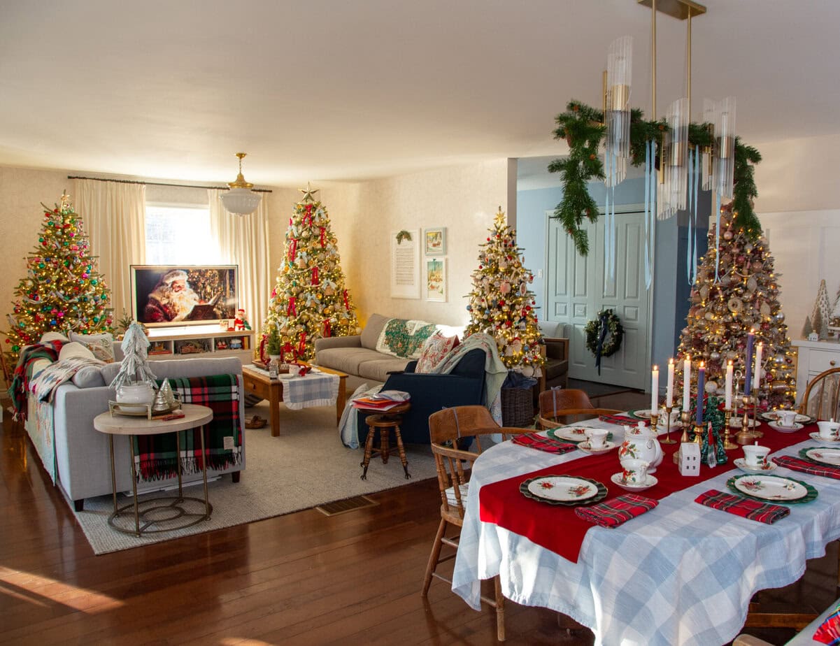 Open-concept living and dining room decorated with multiple Christmas trees, a festive table setting, and warm twinkle lights.