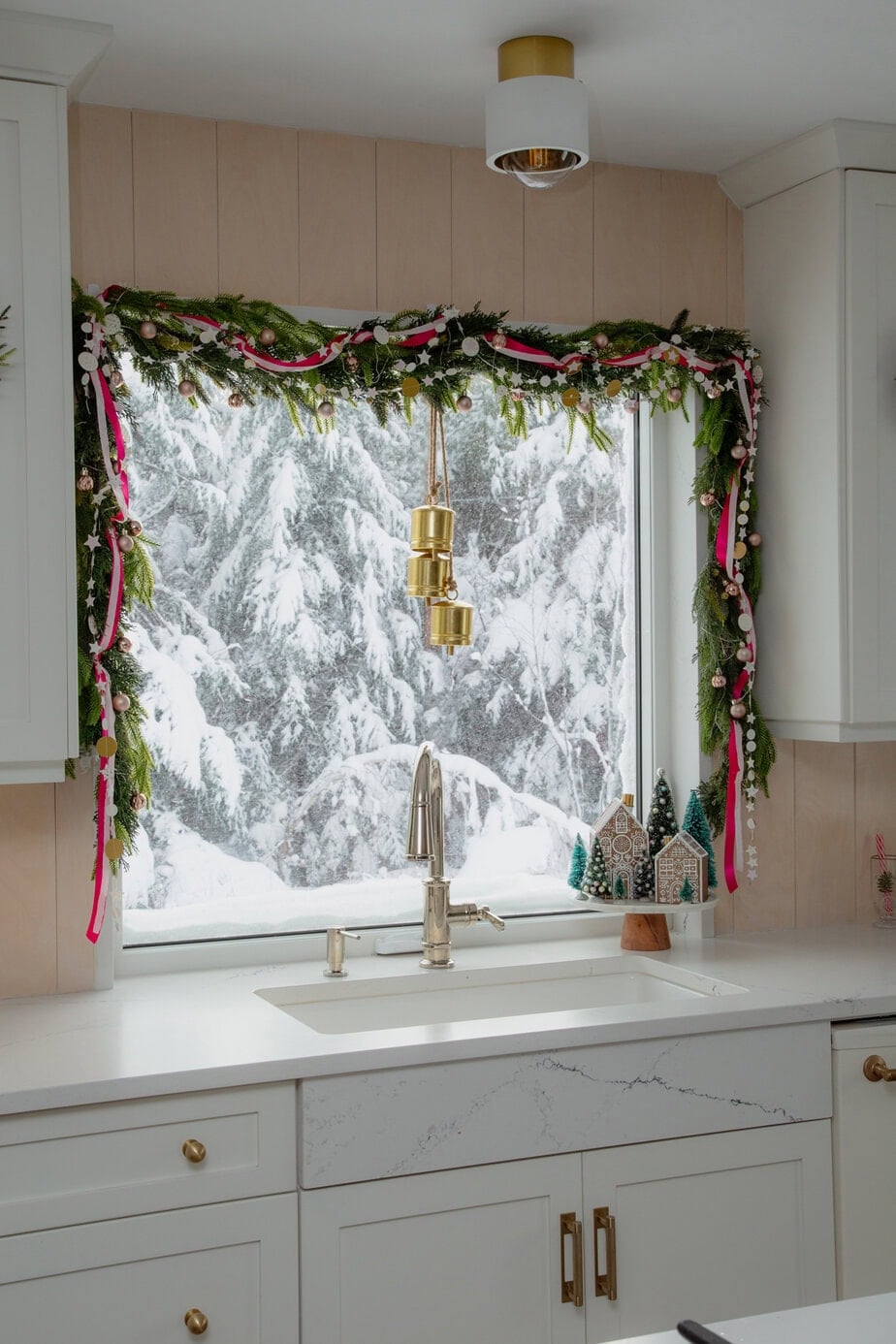Kitchen window dressed with evergreen garland, pink ribbon, and ornaments, with snowy trees outside and a gingerbread village on the sill.
