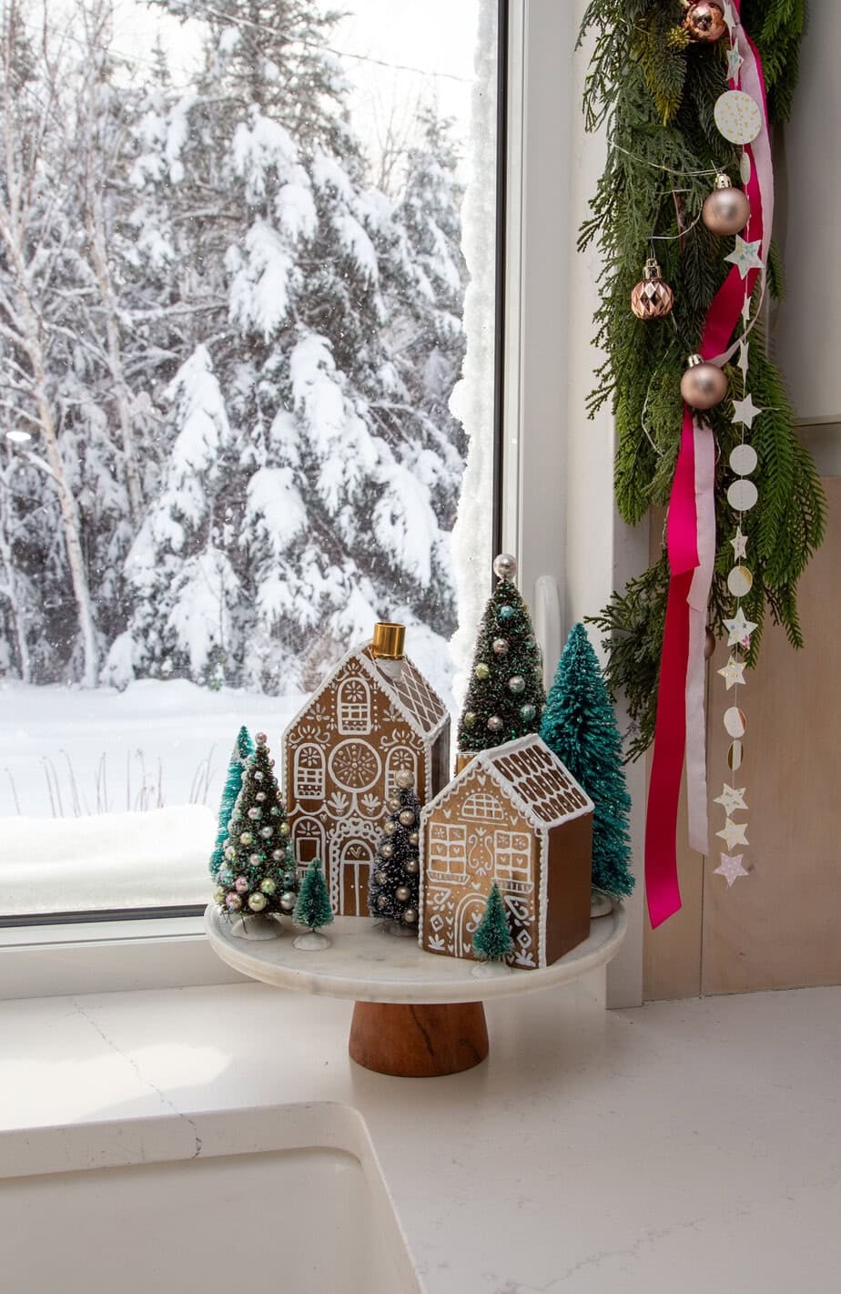 Kitchen window dressed with evergreen garland, pink ribbon, and ornaments, with snowy trees outside and a gingerbread village on the sill.