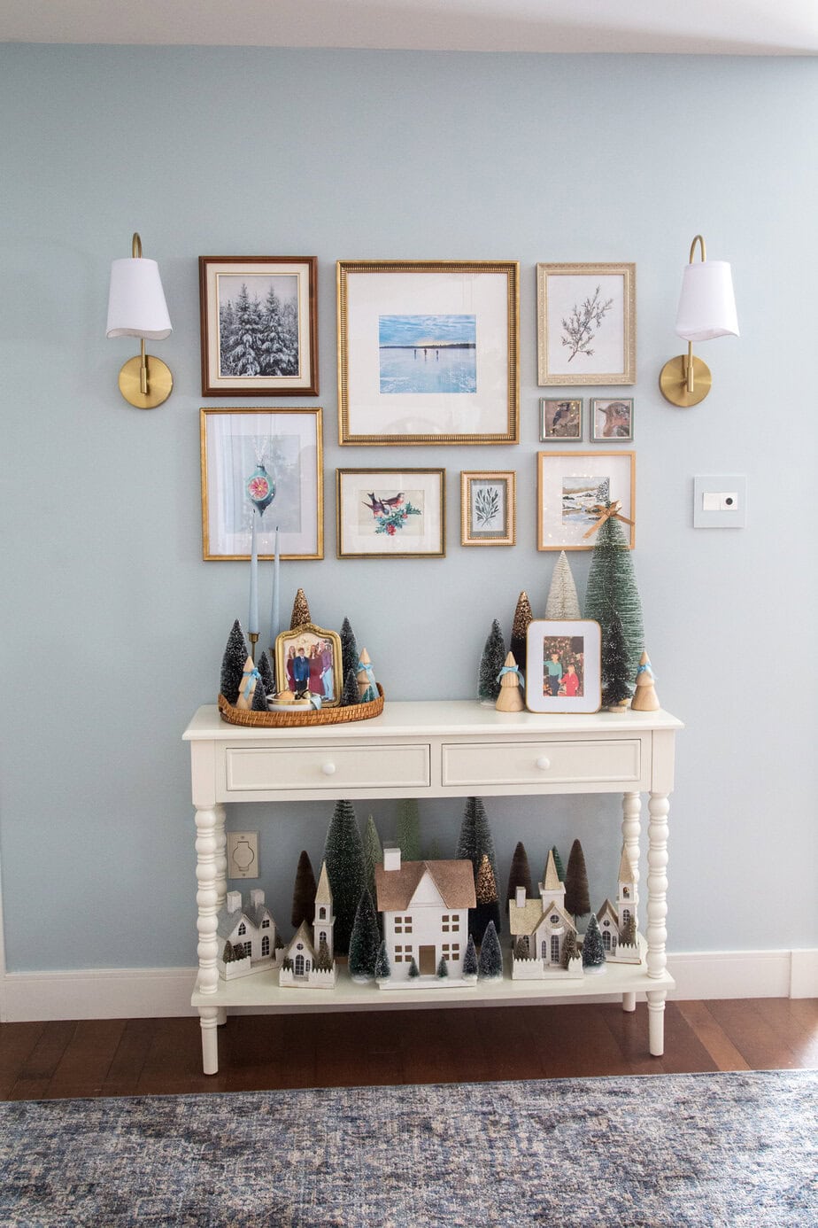 Vintage Christmas console table styled with bottle brush trees, a miniature village, framed family photos, and a winter gallery wall with brass sconces.