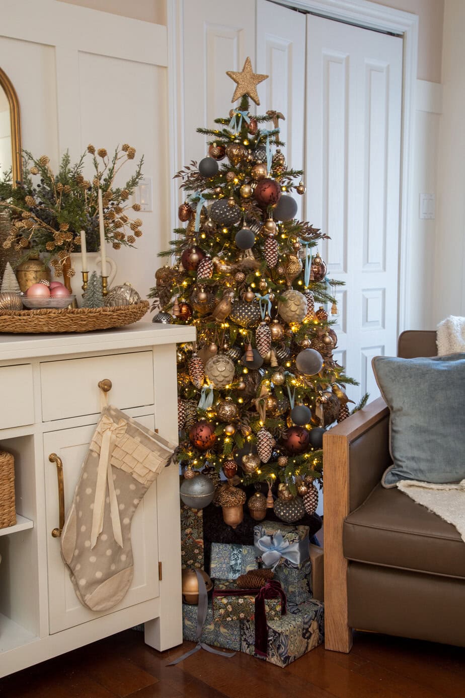 Small vintage Christmas tree with bronze and blue ornaments and a gold star topper beside a white cabinet, stocking, and wrapped gifts.