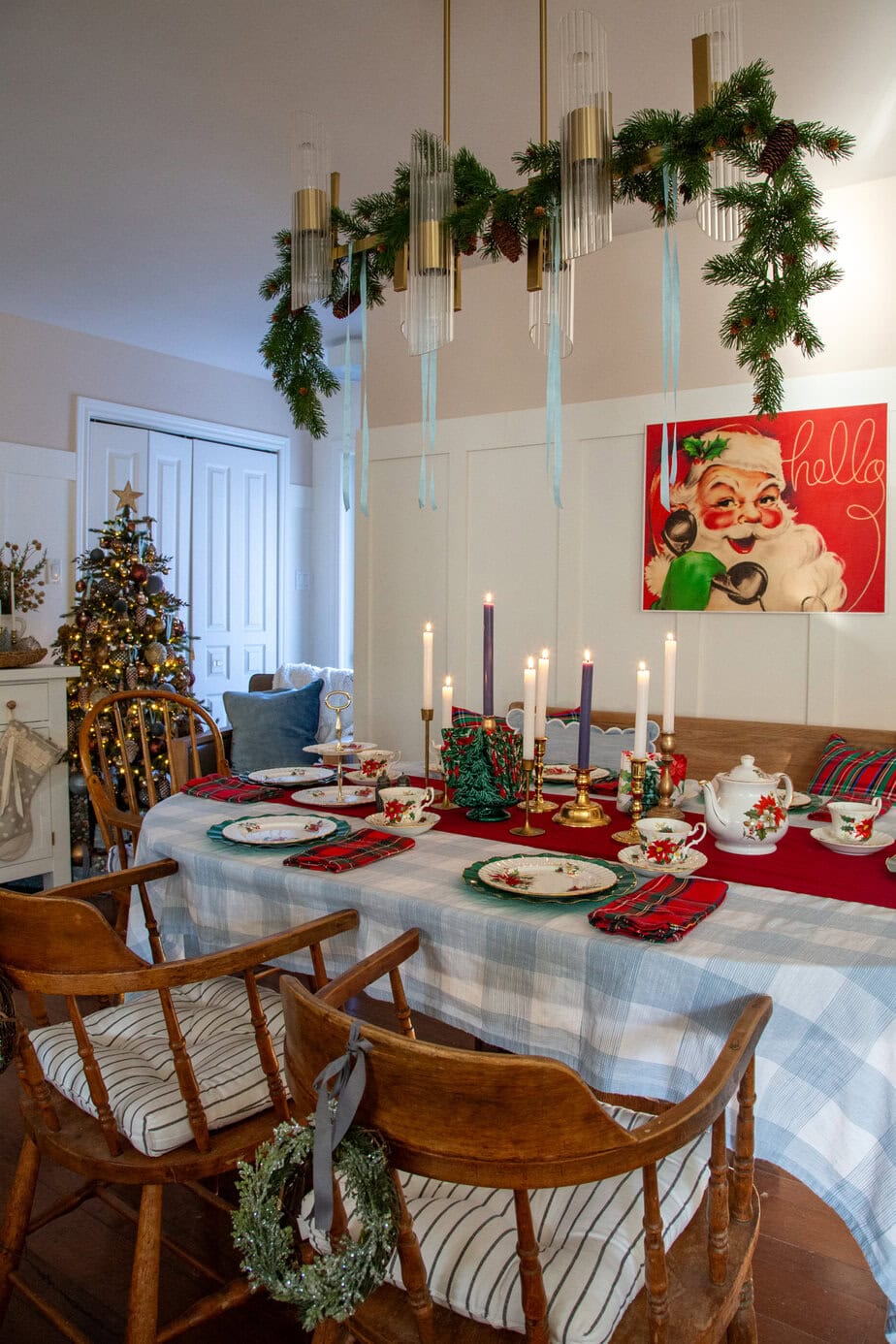 Small vintage Christmas tree with bronze and blue ornaments and a gold star topper beside a white cabinet, stocking, and wrapped gifts. next to dining room table decored for the holidays