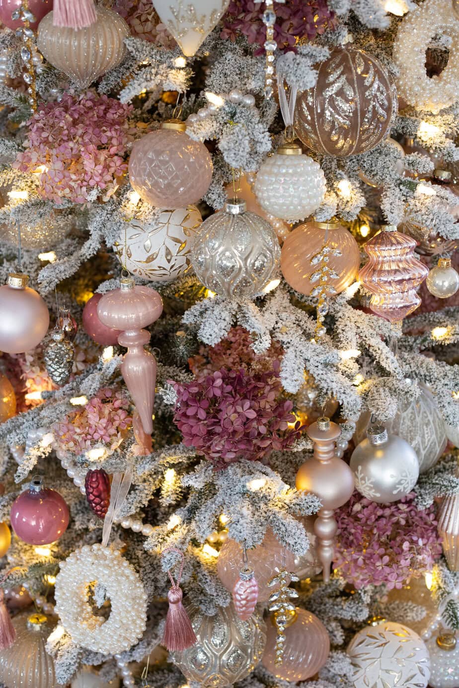 Pink and champagne Christmas tree with hydrangea picks, metallic ornaments, and warm lights in a cozy holiday corner.